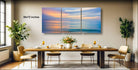 a modern dining room with a large wooden table set for four, surrounded by four chairs. Above the table, there is a large, colorful painting depicting a serene ocean scene with a sunset. The room is decorated with potted plants and other furnishings, creating a warm and inviting atmosphere.