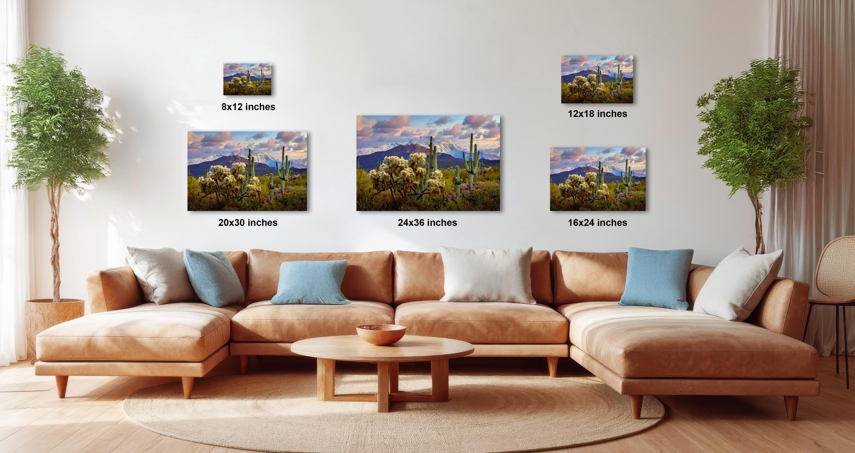 The image is a modern living room with a large sectional sofa, a coffee table, and several potted plants. The wall is decorated with three framed landscape paintings, each varying in size and depicting different scenes. The paintings are arranged in a grid-like pattern, with the largest one at the top and the smallest at the bottom. The room has a warm and inviting atmosphere, with the furniture and decor complementing the overall design.