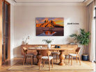 a large, framed photograph of a desert landscape with a sunset, hanging on a wall above a wooden dining table with chairs.
