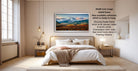 The image is a modern bedroom with a large bed, wooden furniture, and a framed landscape painting hanging on the wall. The painting depicts a mountainous landscape with a sunset sky. The room has a minimalist and clean design, with a white color scheme and natural light coming in from a window.