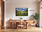 a large landscape painting hanging on a wall in a modern dining room. The painting depicts a serene scene of a lake surrounded by mountains, with a cloudy sky above. The dining table is set with chairs and a vase of flowers, creating a cozy and inviting atmosphere.
