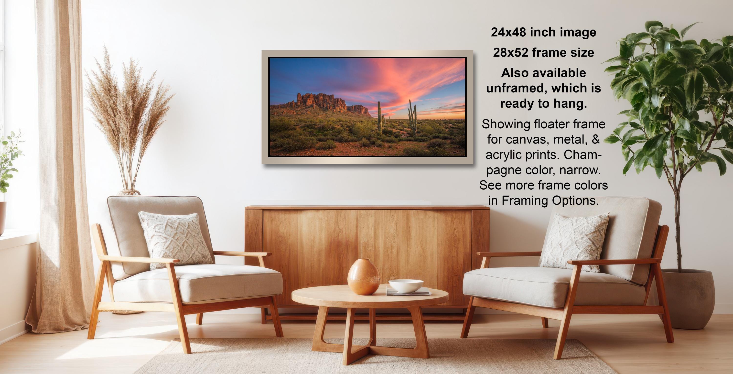 a living room with a large framed landscape painting hanging on the wall. The painting depicts a desert scene with mountains in the background, creating a serene and peaceful atmosphere. The room is furnished with two chairs and a coffee table, providing a comfortable space for relaxation and conversation. The painting's frame size is 24x48 inches, and it is available in a 28x52 frame size. The painting also showcases a floating frame, which adds a modern touch to the overall design.