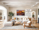 a large, modern living room with a comfortable sofa, a coffee table, and a fireplace. The room has a neutral color scheme with white walls and a gray rug. A large framed landscape painting depicting a desert scene with a castle in the background is hanging on the wall.