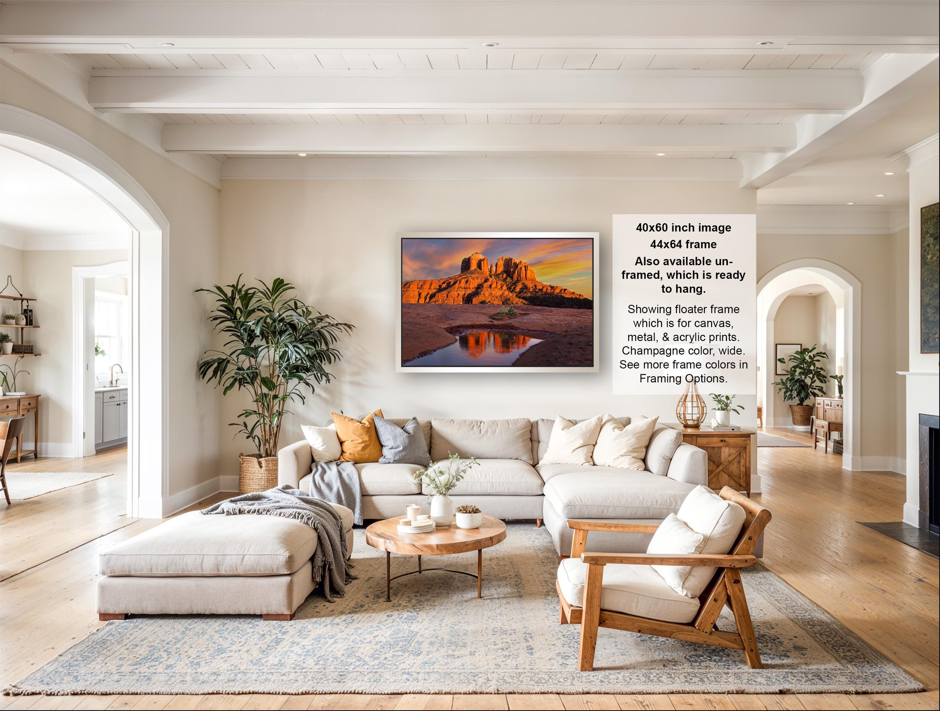 a large, modern living room with a comfortable sofa, a coffee table, and a fireplace. The room has a neutral color scheme with white walls and a gray rug. A large framed landscape painting depicting a desert scene with a castle in the background is hanging on the wall.