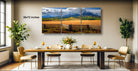 The image is a modern dining room with a large wooden table set for four, surrounded by four chairs. Above the table, there is a large painting depicting a scenic mountain landscape with a cloudy sky. The painting is framed and hangs on the wall, adding a touch of nature and beauty to the room.