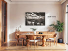 a dining room with a wooden table and chairs, a large framed landscape photograph hanging on the wall, and a potted plant on the right side of the room.