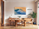a modern dining room with a large wooden table surrounded by matching chairs. A large painting of a crashing wave hangs above the table, adding a dramatic element to the space. The room is decorated with various plants and other decorative items, creating a cozy and inviting atmosphere.
