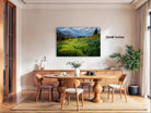 a serene dining room with a large wooden table surrounded by matching chairs. A painting of a lush green field with yellow flowers hangs on the wall above the table. The room is decorated with potted plants and other natural elements, creating a peaceful and inviting atmosphere.