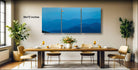 a modern dining room with a large wooden table set for four, surrounded by four chairs. Above the table, there is a large canvas print depicting a serene mountain landscape with a bright orange sun in the sky. The room is decorated with potted plants and other furnishings, creating a cozy and inviting atmosphere.