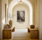 a hallway with a wooden floor, arched doorways, and two wooden benches. There is a large black and white photograph hanging on the wall, and a vase with flowers on one of the benches. The hallway has a warm, inviting atmosphere with natural light coming in from the windows.
