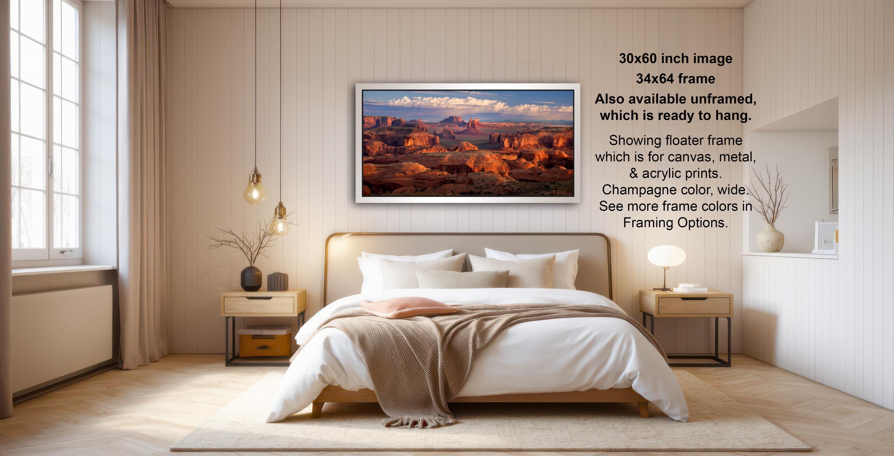 The image is a bedroom with a large bed, a nightstand, and a painting of a desert landscape hanging on the wall. The painting is framed in a 34x64-inch frame, which is available unframed, and it is ready to hang. The bedroom has a minimalist and modern design, with a neutral color palette and a focus on the bed and nightstand.
