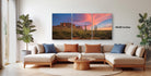 a living room with a large, colorful landscape painting hanging on the wall. The painting depicts a desert scene with mountains and a sunset sky. The room has a comfortable seating arrangement with a sofa and a coffee table. There are also several potted plants and decorative items in the room, creating a cozy and inviting atmosphere.