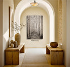 a hallway with wooden benches and tables, a large framed picture of a forest on the wall, and a vase of flowers on one of the tables.