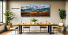 a large, three-part landscape painting depicting a mountainous, mountainous region with a cloudy sky. The painting is hung on a wall above a dining table set with wine glasses, plates, and a vase of flowers.