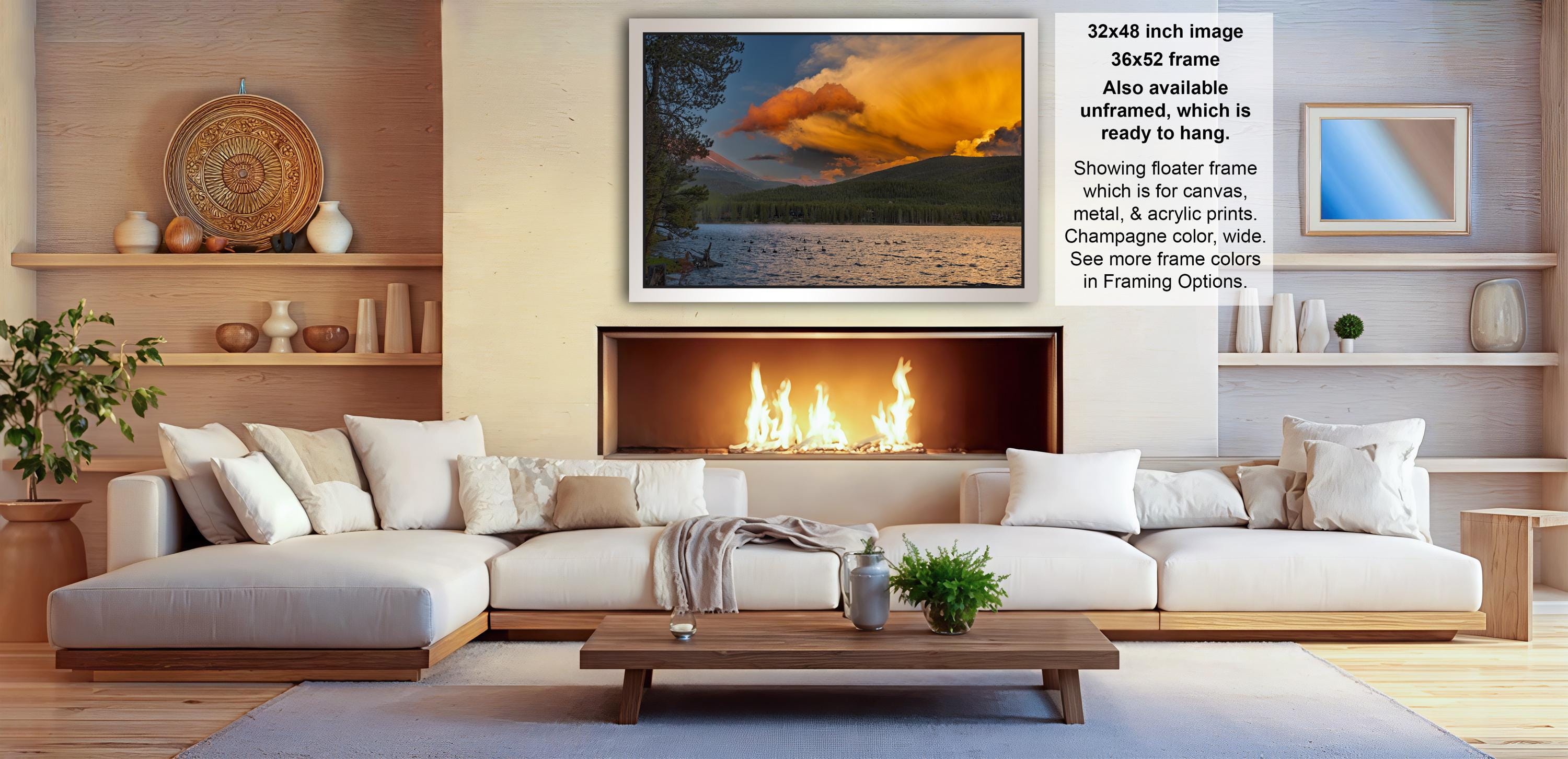 a cozy living room with a fireplace, white sofas, and a coffee table. The room has a warm and inviting atmosphere, with a large framed picture of a sunset over a lake hanging on the wall.