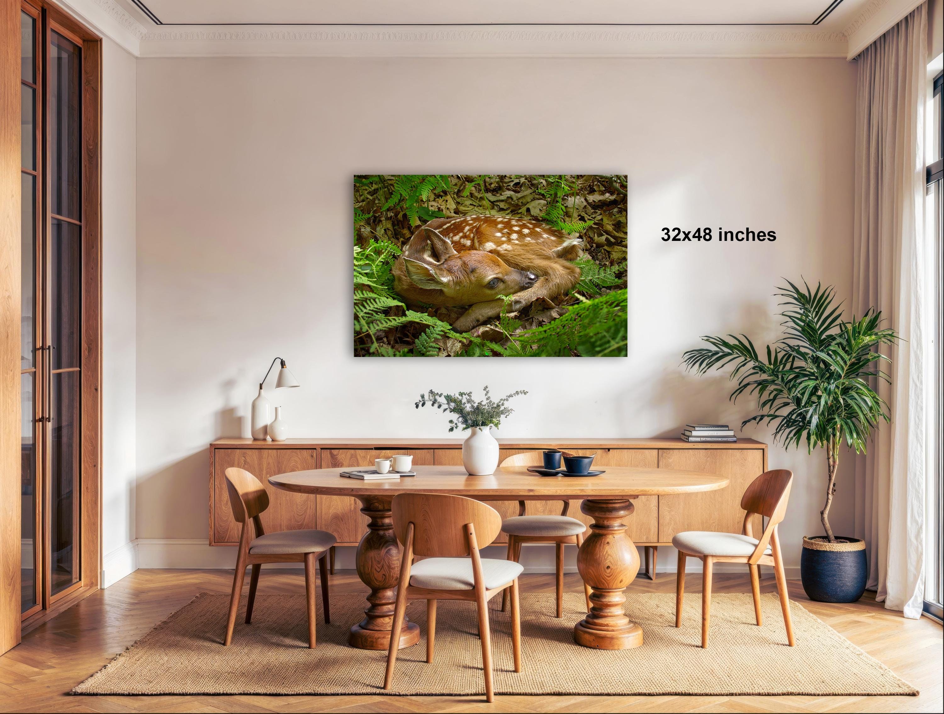 a serene indoor setting with a dining table surrounded by chairs, a large framed photograph of a deer resting in the forest, and a potted plant on the right side of the room.
