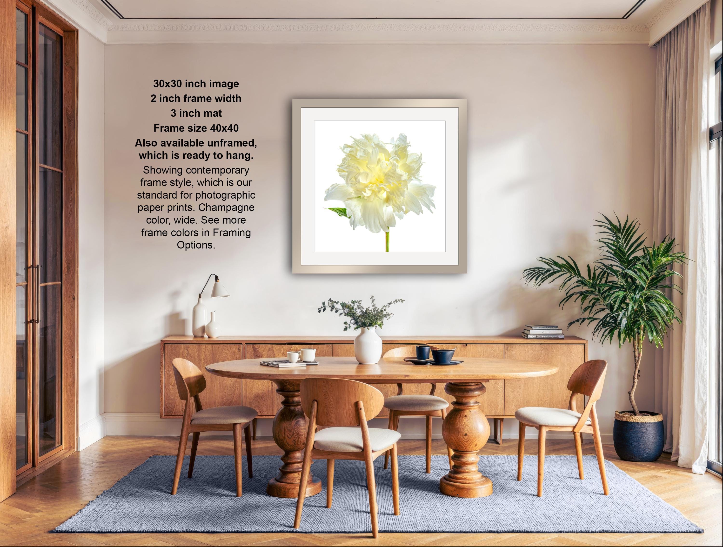 The image is a modern living room with a large white flower in a frame hanging on the wall. The room has a wooden dining table surrounded by chairs, and there are potted plants and other decorative items in the space.