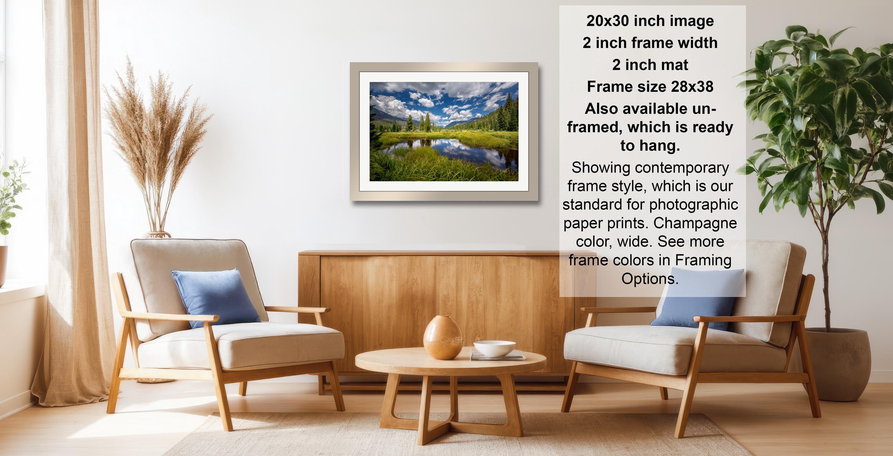 The image is a living room with a framed landscape photograph hanging on the wall, a wooden coffee table in the center, and two chairs facing each other.