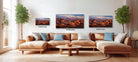 a large, brown sectional sofa with three framed landscape paintings hanging above it. The paintings depict desert scenes with mountains in the background. The sofa is positioned in front of a window, and there are two potted plants on either side of the sofa. The paintings are of different sizes, with the largest one being 24 inches wide and the smallest one being 12 inches wide.