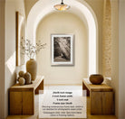 The image is a photograph of a hallway with a framed black and white photograph hanging on the wall, flanked by two wooden tables with decorative items on them.
