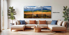 The image is a modern living room with a large, three-panel landscape painting hanging on the wall. The painting depicts a scenic countryside with mountains in the background and a field in the foreground. The room is furnished with a sofa and a coffee table, creating a comfortable and inviting atmosphere.