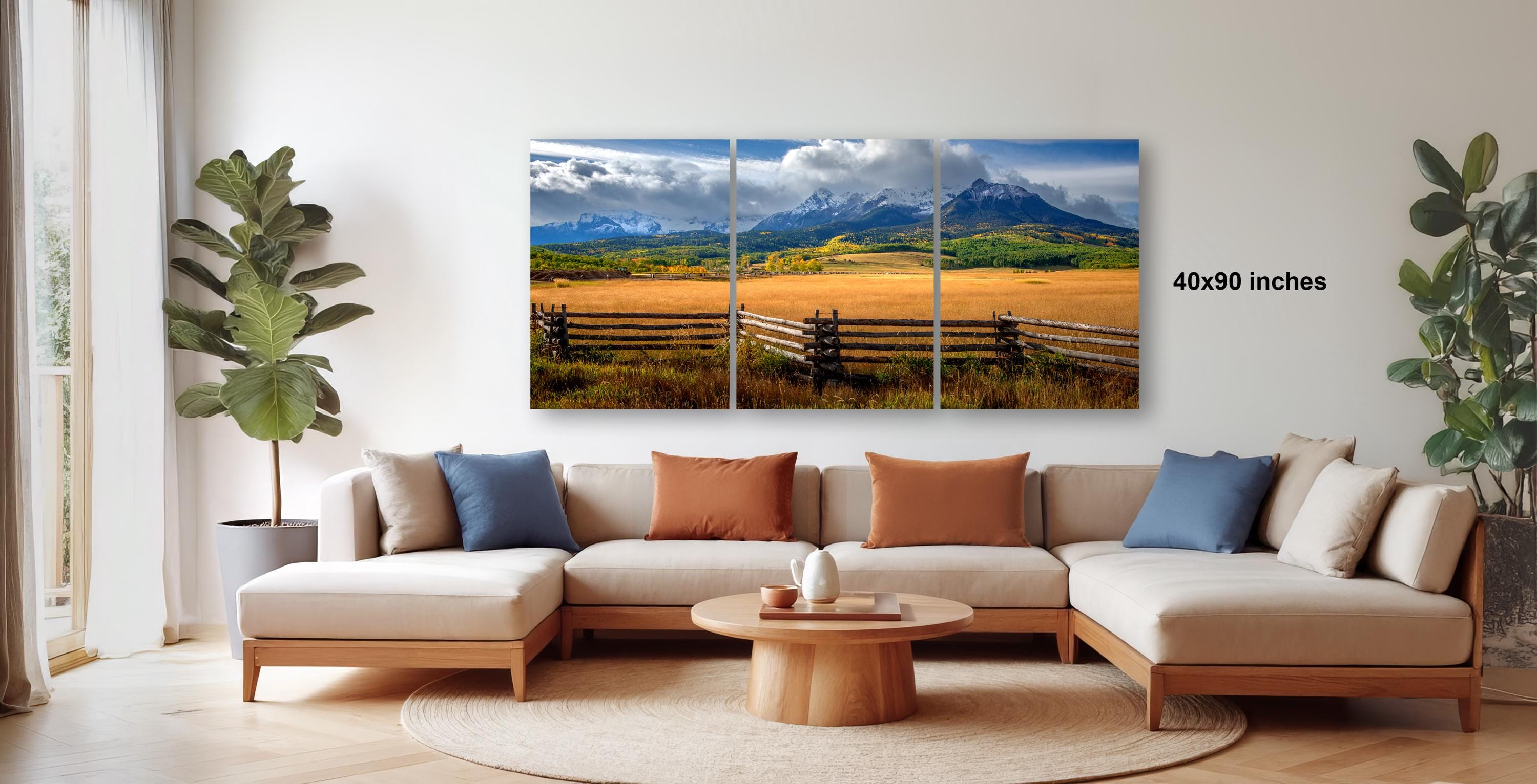 The image is a modern living room with a large, three-panel landscape painting hanging on the wall. The painting depicts a scenic countryside with mountains in the background and a field in the foreground. The room is furnished with a sofa and a coffee table, creating a comfortable and inviting atmosphere.