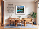 The image is a modern living room with a large framed photograph of a scenic mountain landscape hanging on the wall. The photograph is framed in a 2x4-inch frame with a 3-inch mat, and it is ready to be hung. The room has a wooden dining table surrounded by chairs, and there are potted plants and other decorative items on the table.