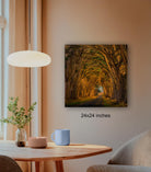 a cozy indoor setting with a wooden table, a lamp, a blue mug, and a plant. Above the table, there is a framed photograph of a tree-lined path leading to a tunnel, creating a sense of depth and mystery.