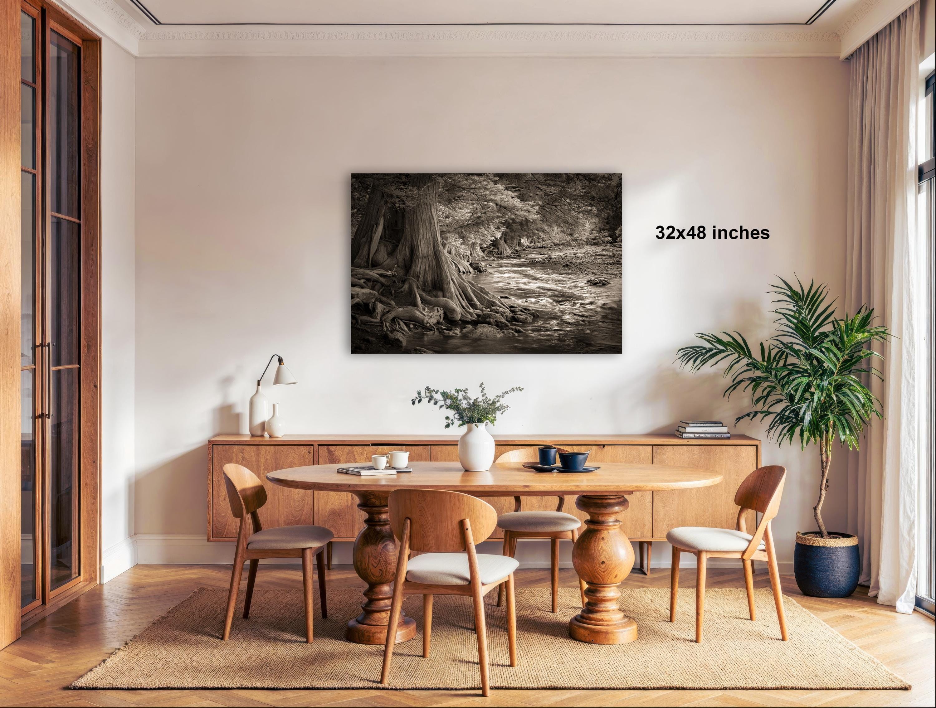 a cozy dining room with a wooden table and chairs, a large framed black and white photograph of a forest scene hanging on the wall, and a potted plant and vase on the table.