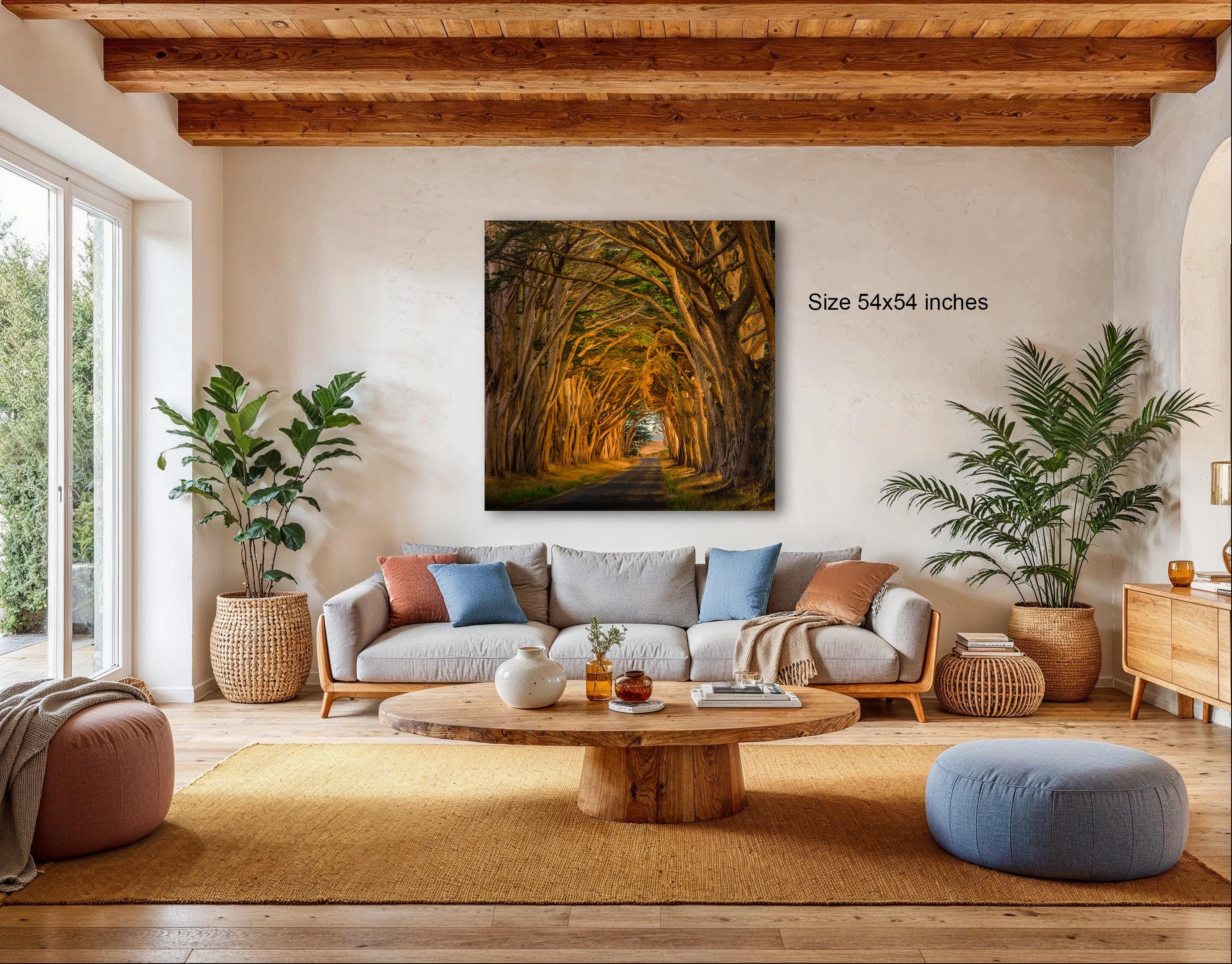 a cozy living room with a large window overlooking a tree-lined path. The room is furnished with a gray sofa, a wooden coffee table, and several potted plants. A large framed painting of a tree-lined path hangs on the wall, adding a natural element to the space.