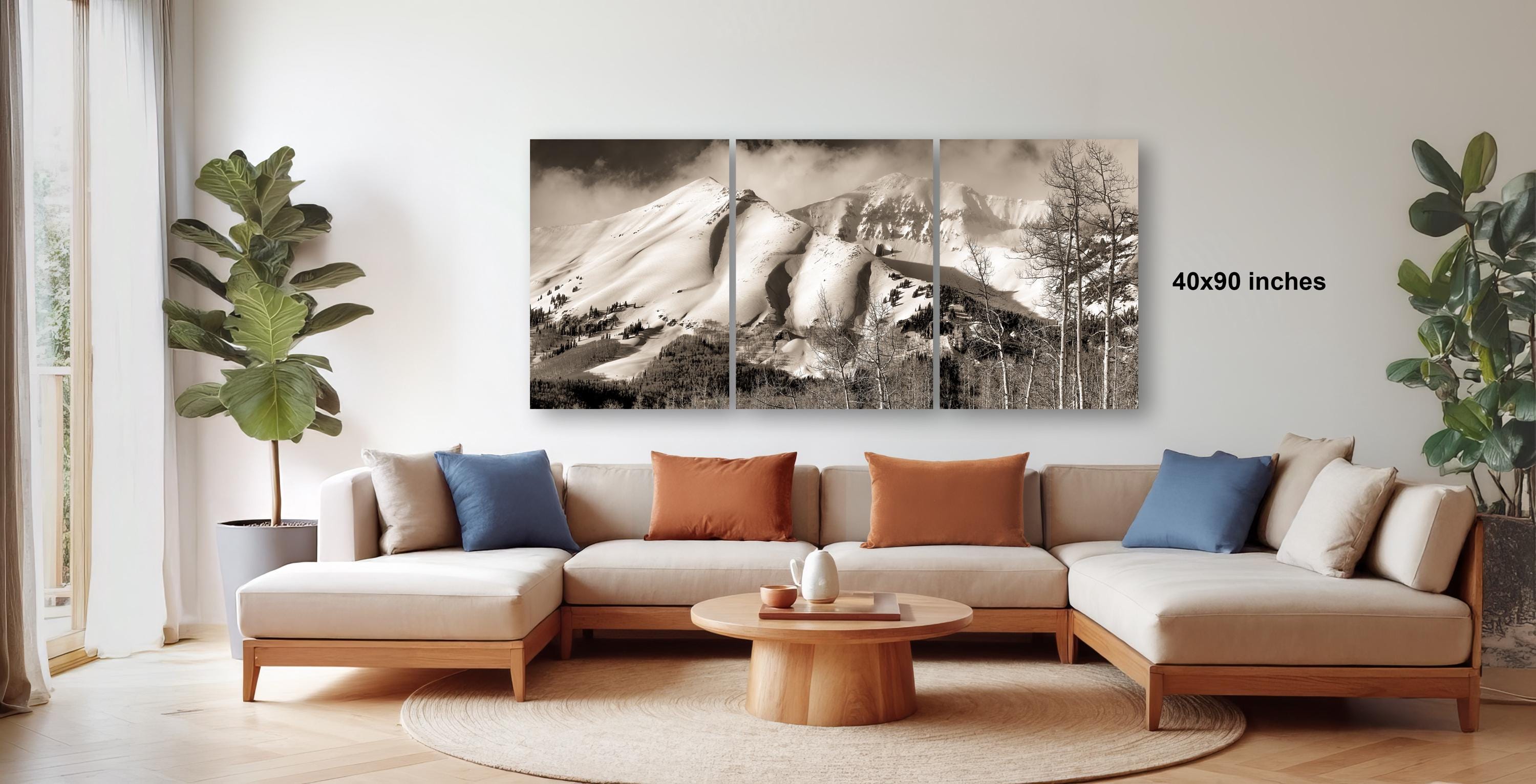 a modern living room with a large, black and white landscape photograph of a mountain range hanging on the wall. The photograph is composed of three panels, and the text "4090 inches" is visible, indicating the size of the artwork. The room is furnished with a beige sofa and a wooden coffee table, creating a cozy and inviting atmosphere.