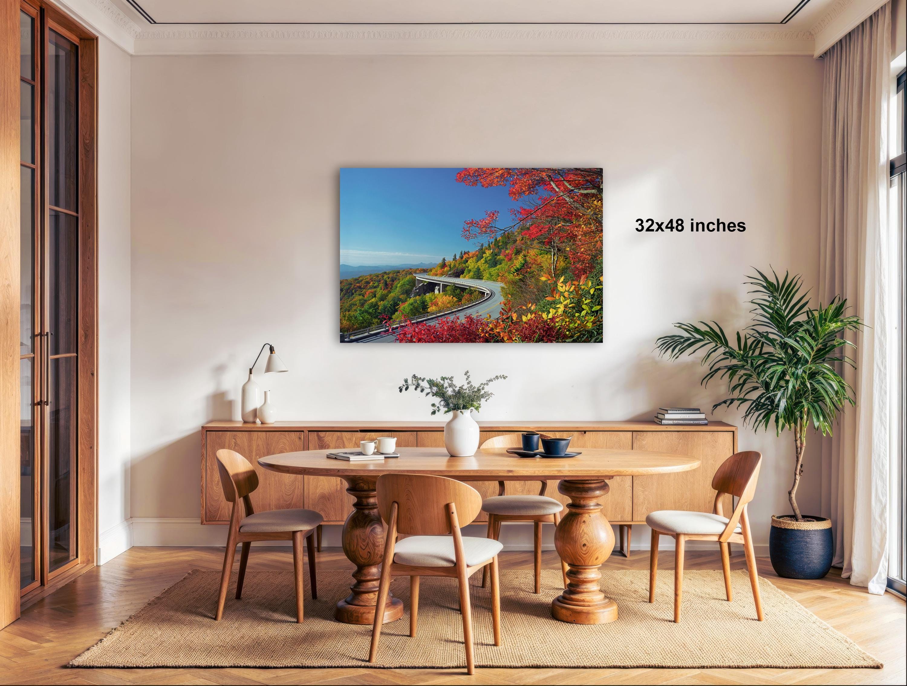 a dining room with a large wooden table surrounded by chairs, a vase of flowers, and a painting of a scenic mountain road.
