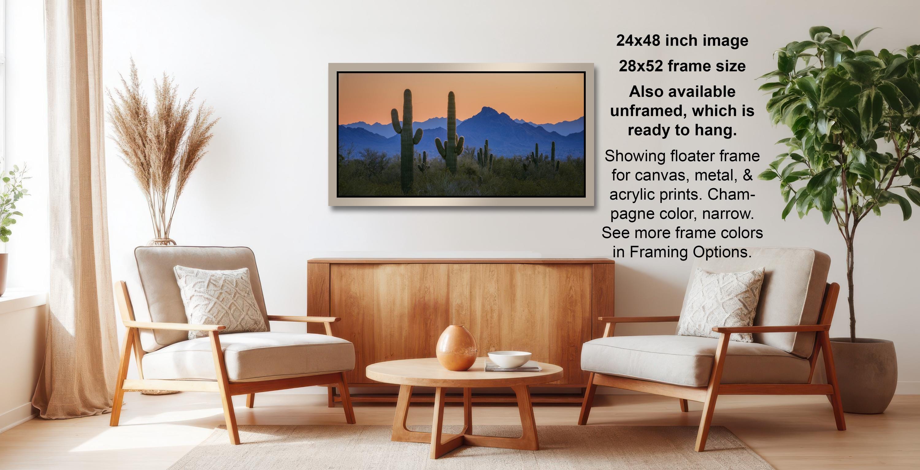 a living room with a wooden coffee table, two white armchairs, and a framed landscape painting of a desert scene with cacti and mountains in the background.