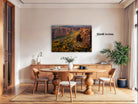 a large, framed landscape painting hanging on a wall above a wooden dining table with chairs.