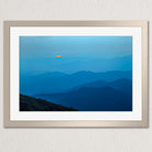 A framed photograph depicting a breathtaking mountain landscape with a vibrant orange sun peeking through the hazy blue mountains.