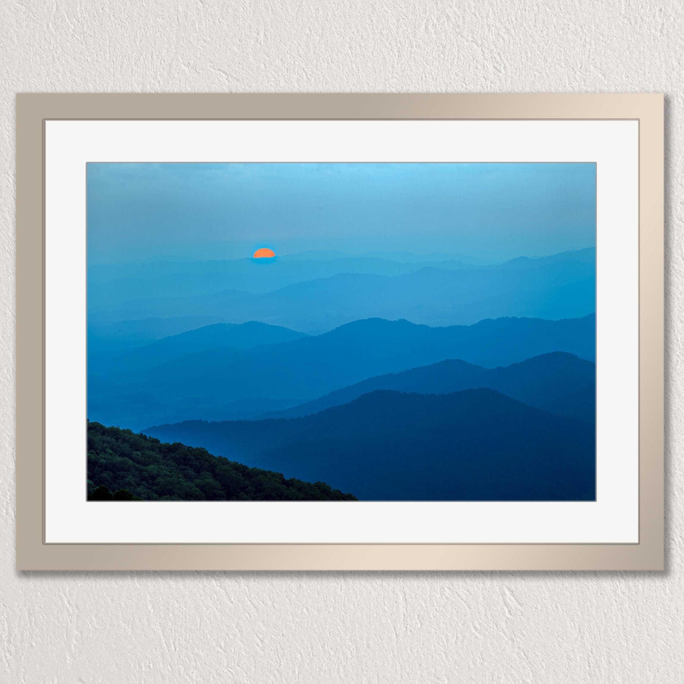 A framed photograph depicting a breathtaking mountain landscape with a vibrant orange sun peeking through the hazy blue mountains.