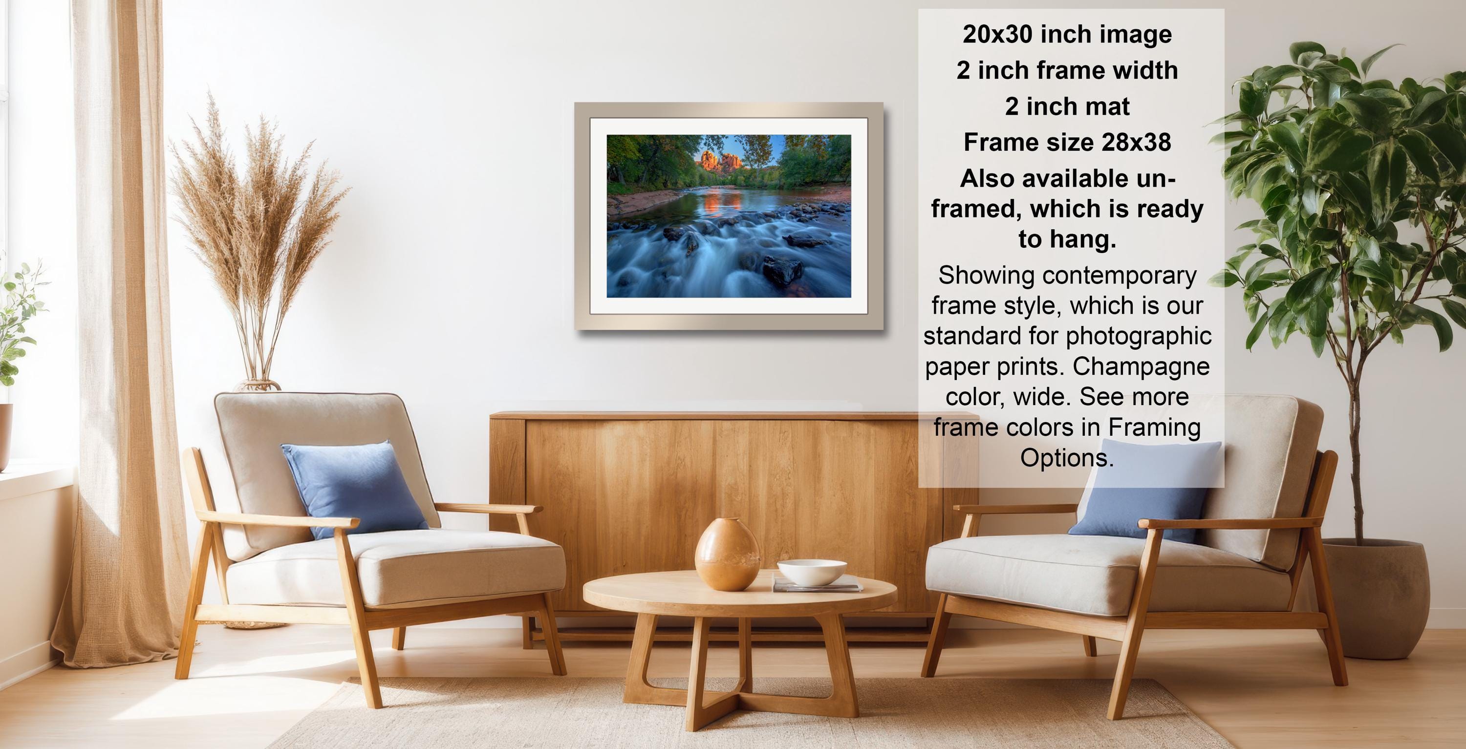 a living room with a framed photograph of a river and waterfall hanging on the wall. The room has two armchairs, a coffee table, and a potted plant. The photograph is framed in a modern style, and the frame size is 28.38 inches wide. The text indicates that the frame is available in both framed and unframed versions, and it is ready to hang.