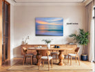 a serene dining room with a large wooden table surrounded by matching chairs. A painting of a sunset is displayed on the wall above the table, creating a warm and inviting atmosphere. The room is decorated with various items, including a potted plant, a vase, and a lamp, adding to the overall ambiance.