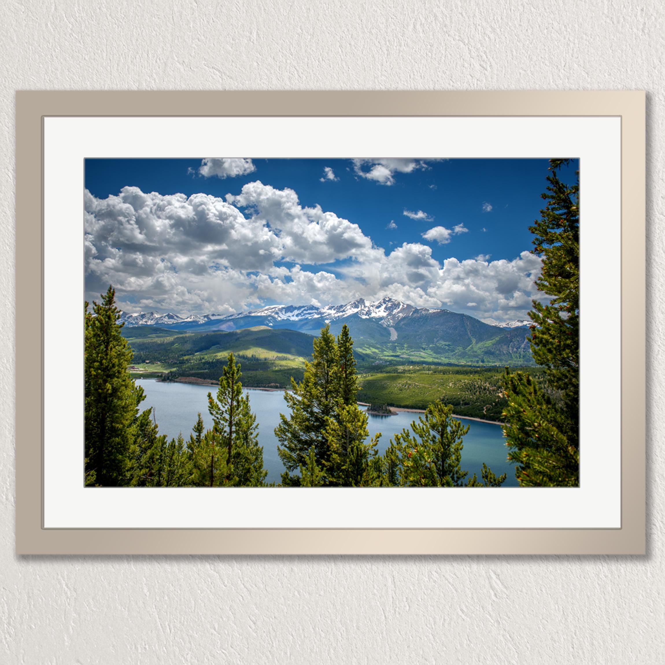 a serene landscape with a lake surrounded by lush green trees and majestic mountains in the distance. The sky is a clear blue with fluffy white clouds, creating a picturesque scene.