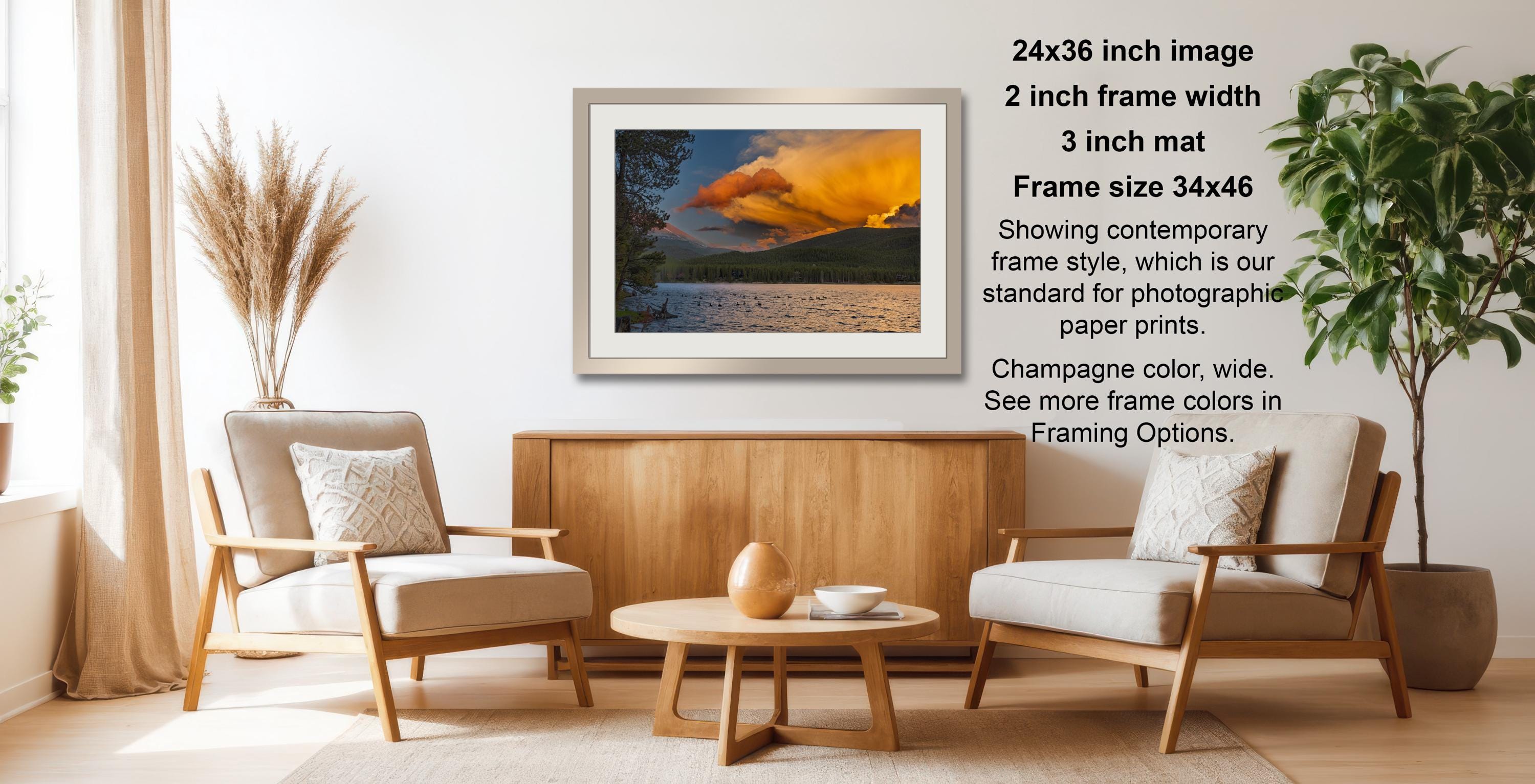 a living room with a wooden coffee table, two white armchairs, and a framed photograph of a sunset over a body of water. The photograph is framed in a 2x3 grid format and is displayed on a white wall.