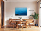 a modern dining room with a large wooden table surrounded by matching chairs. A large painting of a mountain landscape hangs on the wall above the table. The room is decorated with a potted plant, a vase, and a lamp, creating a cozy and inviting atmosphere.