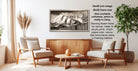 The image is a living room with a wooden coffee table, two white armchairs, and a framed black and white photograph of a mountain landscape hanging on the wall.