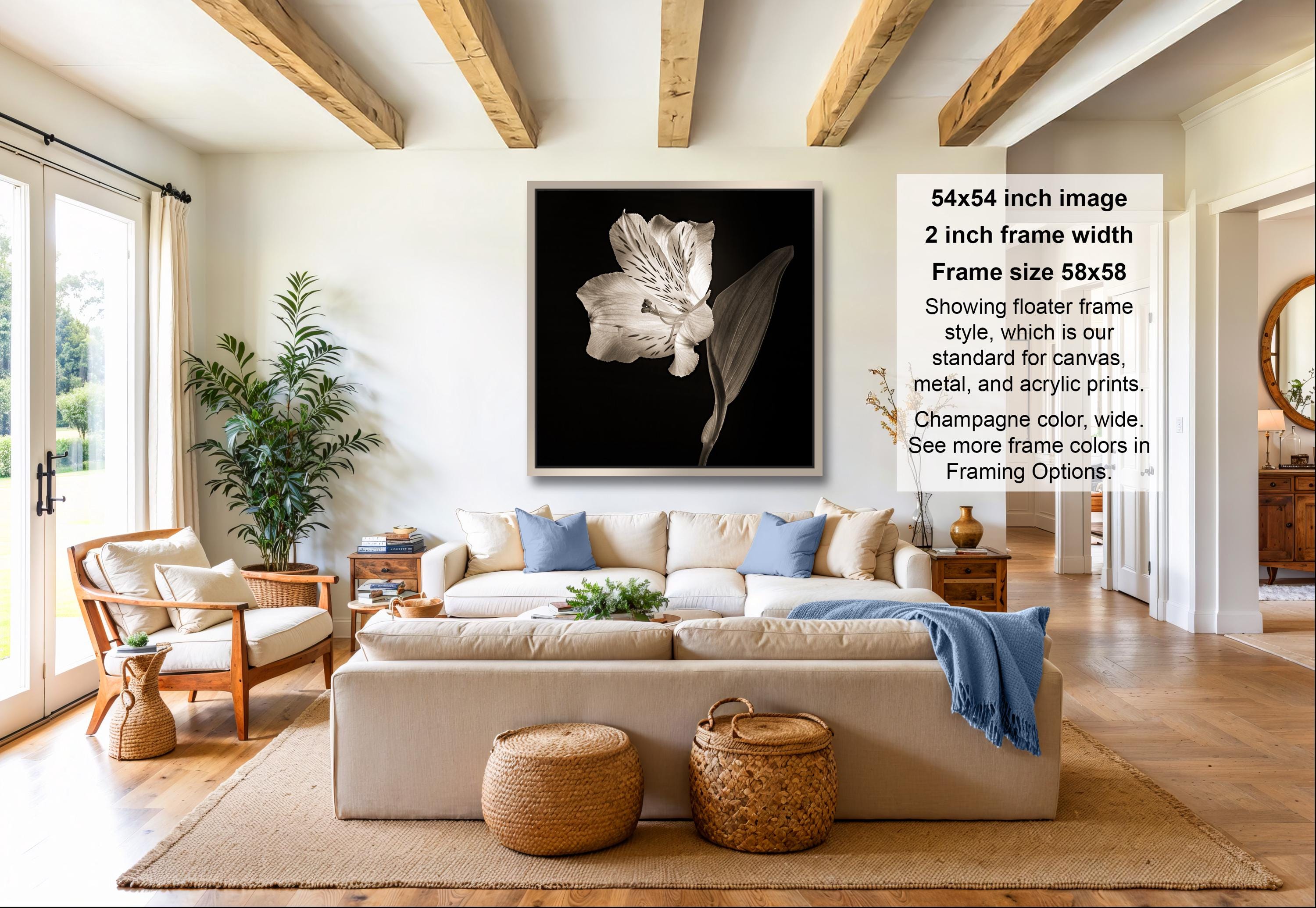 A modern living room with a large white sofa, a wooden coffee table, and a potted plant. The room has a minimalist and clean design, with a large framed black and white photograph of a flower hanging on the wall.