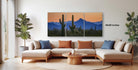a modern living room with a large, three-panel landscape painting of a desert scene hanging on the wall. The painting depicts mountains and cacti in the background, creating a serene and natural atmosphere. The room is furnished with a beige sofa and a wooden coffee table, and the space is decorated with potted plants and other natural elements, contributing to the overall aesthetic.