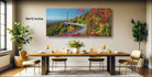 a large, colorful painting of a winding road through a forested mountain landscape, hanging on a wall above a wooden dining table set with wine glasses, plates, and a vase of flowers.