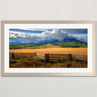 A picturesque rural landscape with a wooden fence in the foreground, a vast field in the middle ground, and majestic mountains in the background under a cloudy sky.