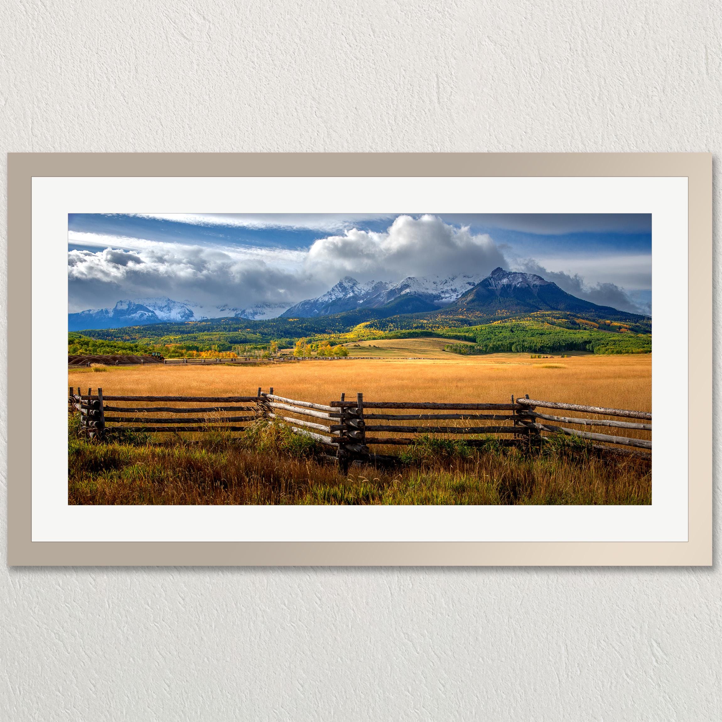 A picturesque rural landscape with a wooden fence in the foreground, a vast field in the middle ground, and majestic mountains in the background under a cloudy sky.
