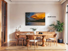 a modern dining room with a large wooden table surrounded by matching chairs. A large painting of a sunset landscape is displayed on the wall above the table. The room is decorated with various plants and other furnishings, creating a cozy and inviting atmosphere.
